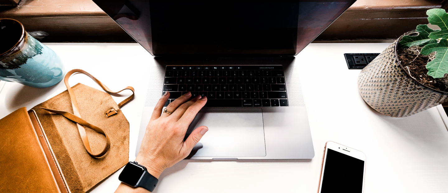 Computer open with hand touching keypad, leather satchel, plant, and coffee mug